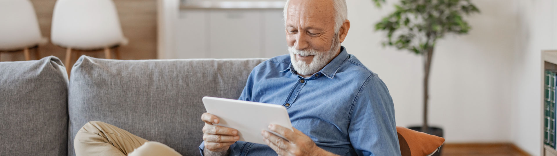 A senior man sitting on a couch while using a tablet.