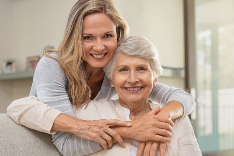 Cheerful mature woman embracing senior mother at home and looking at camera. Portrait of elderly mother and middle aged daughter smiling together. Happy daughter embracing from behind elderly mom.