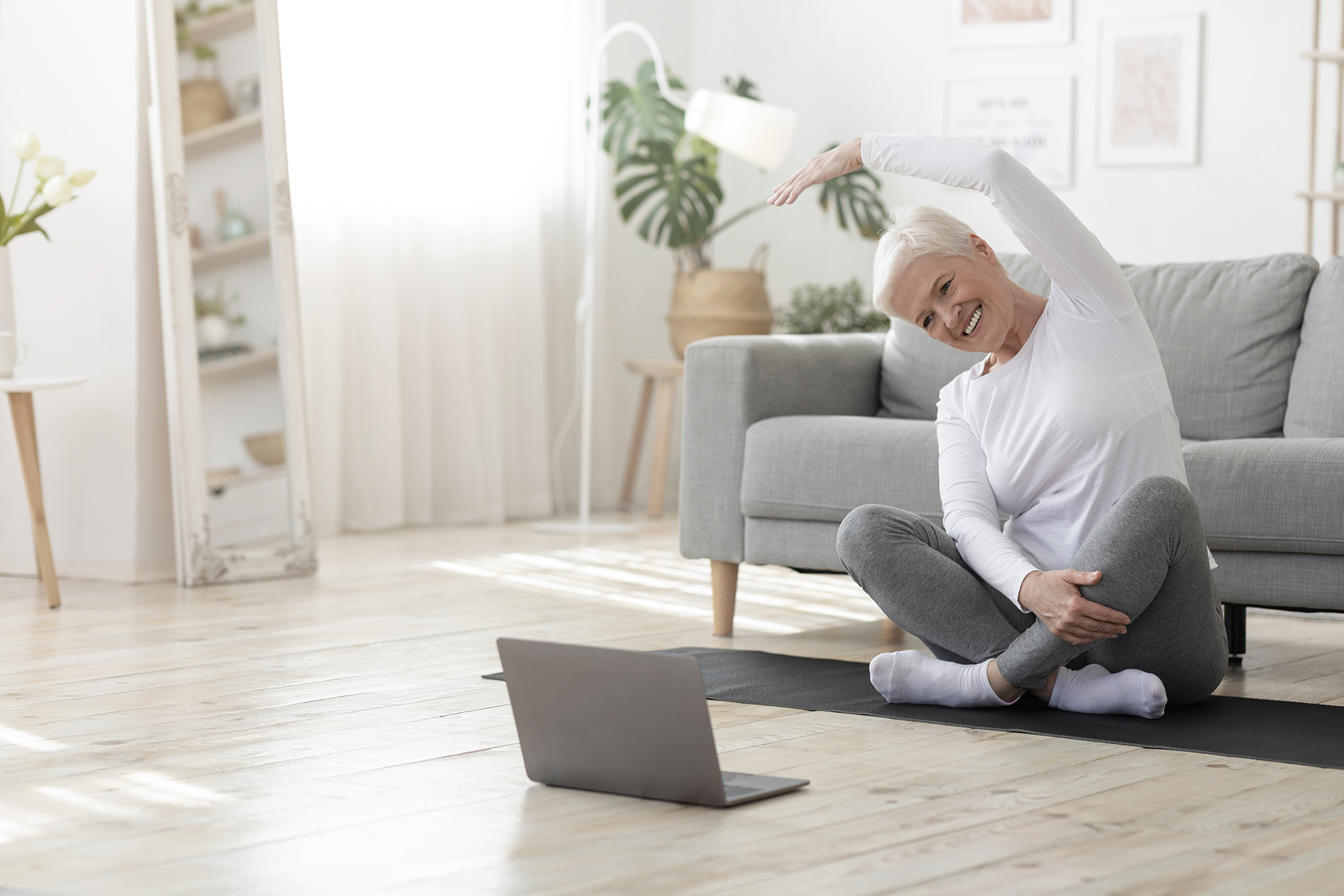 A senior woman practices yoga while watching a virtual class on a laptop.