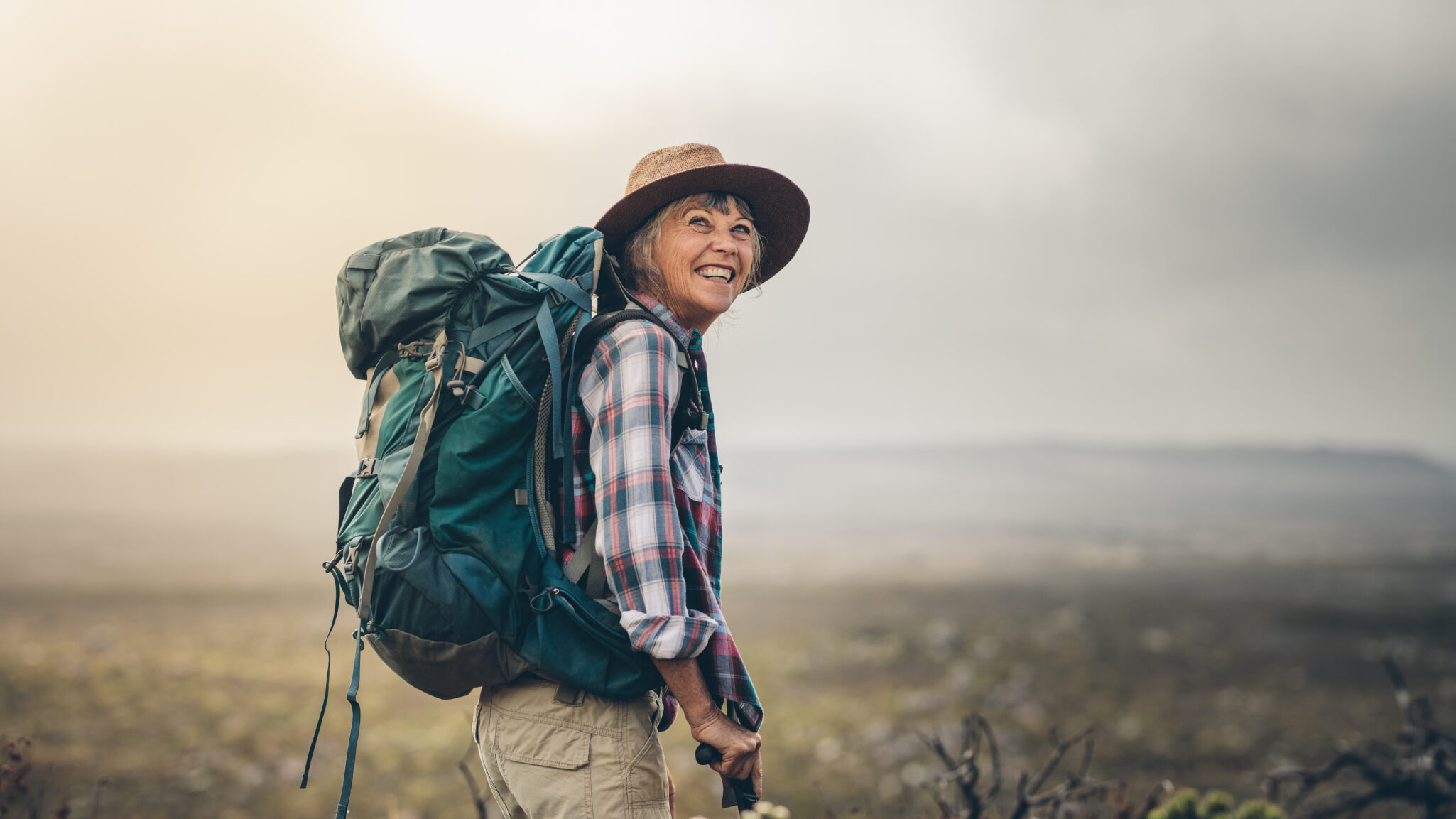 A senior woman stops and smiles while hiking with a pack on her back.