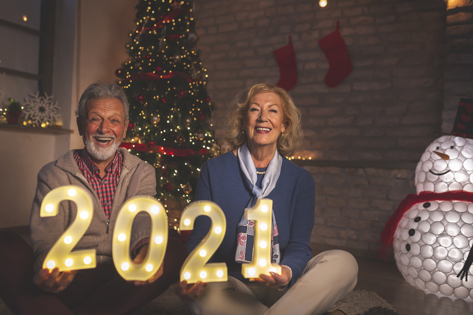 A senior couple pose for a photo in front of a Christmas tree while holding lighted plastic numbers for 2021.