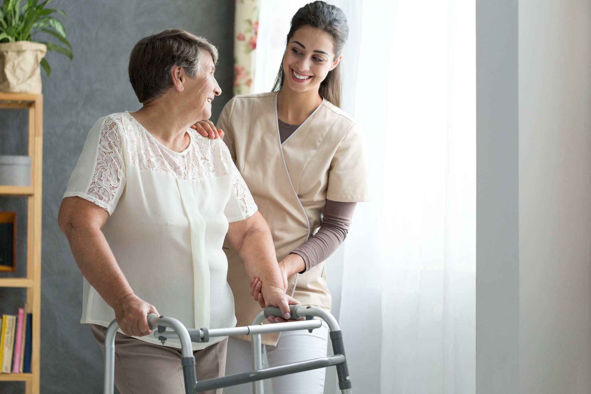 A senior woman walks with assistance from a health care staff member.