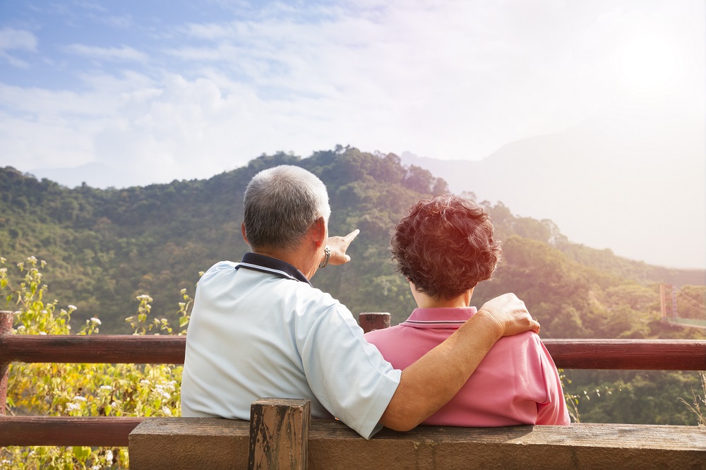 A senior couple sit on a bench and enjoy a scenic view of rolling hills.