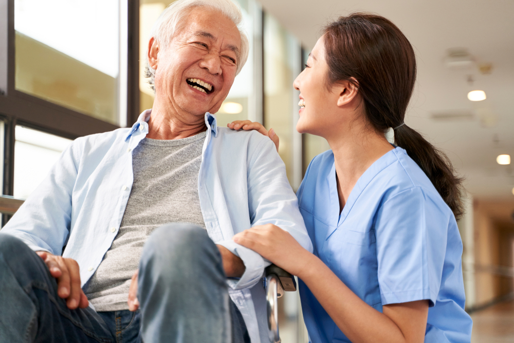 A senior man sitting in a wheelchair laughs with a female nurse.