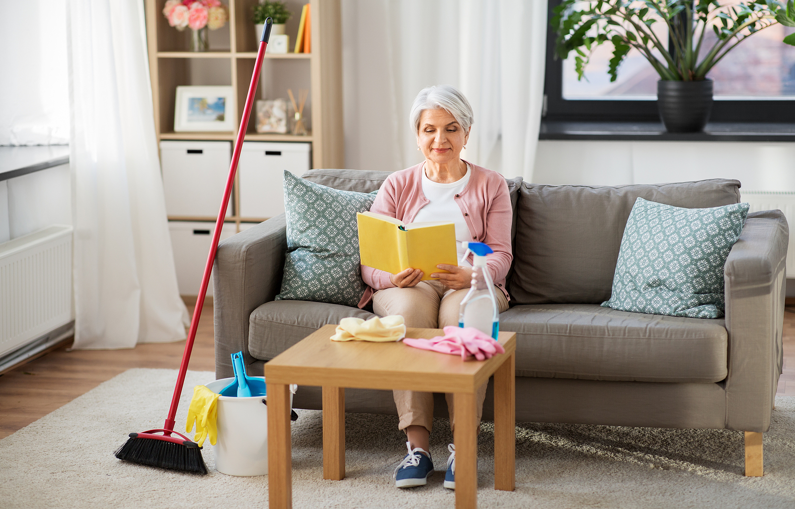 A senior woman sits on a sofa and reads a book with cleaning supplies sitting nearby.