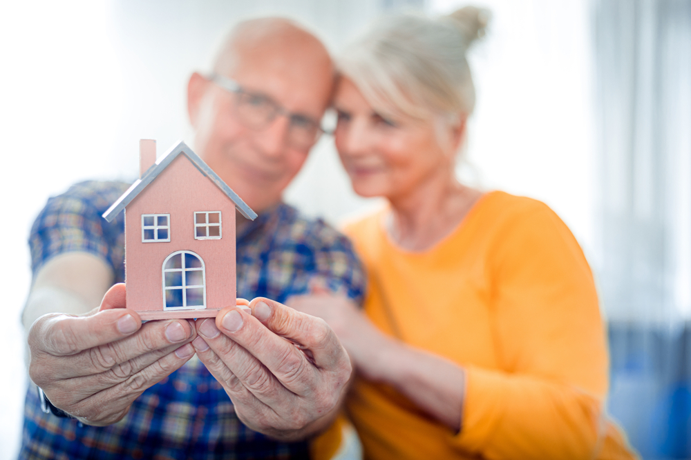 A senior citizen couple touch heads while the man holds a wooden block house out.