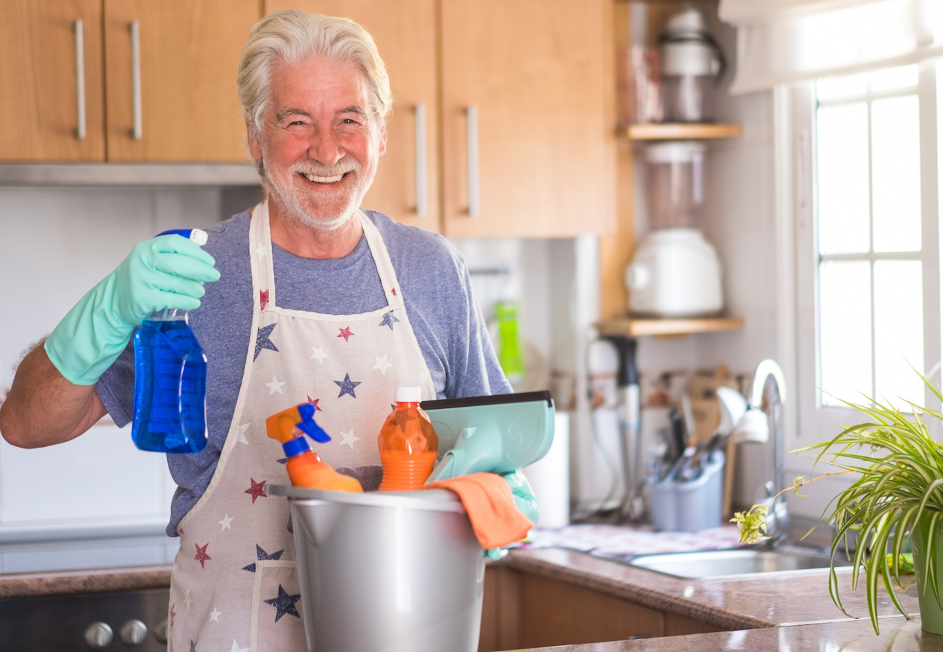 A smiling senior man holds cleaning supplies with gloves on.