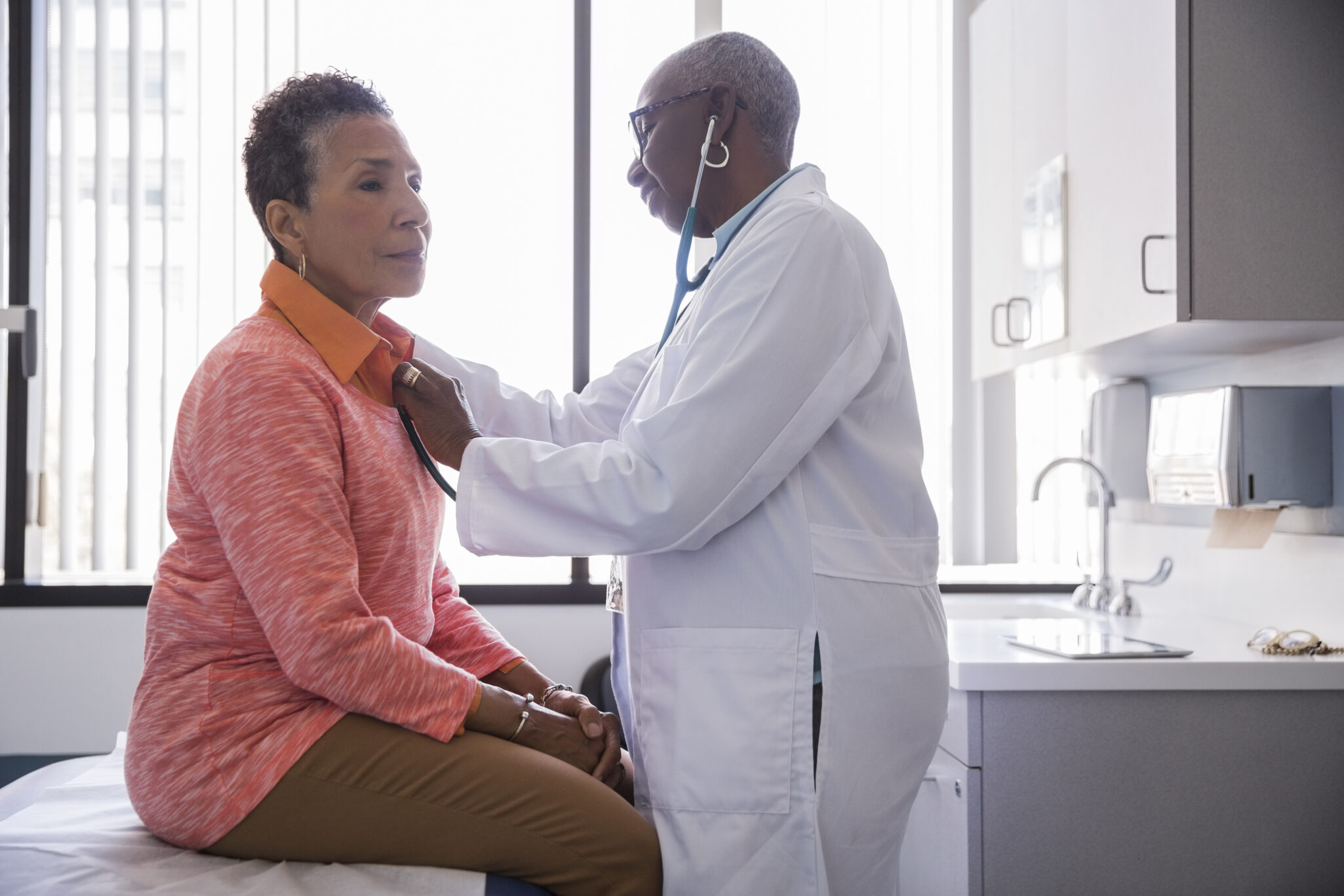 A senior woman sitting on an exam table while a female doctor performs an exam.