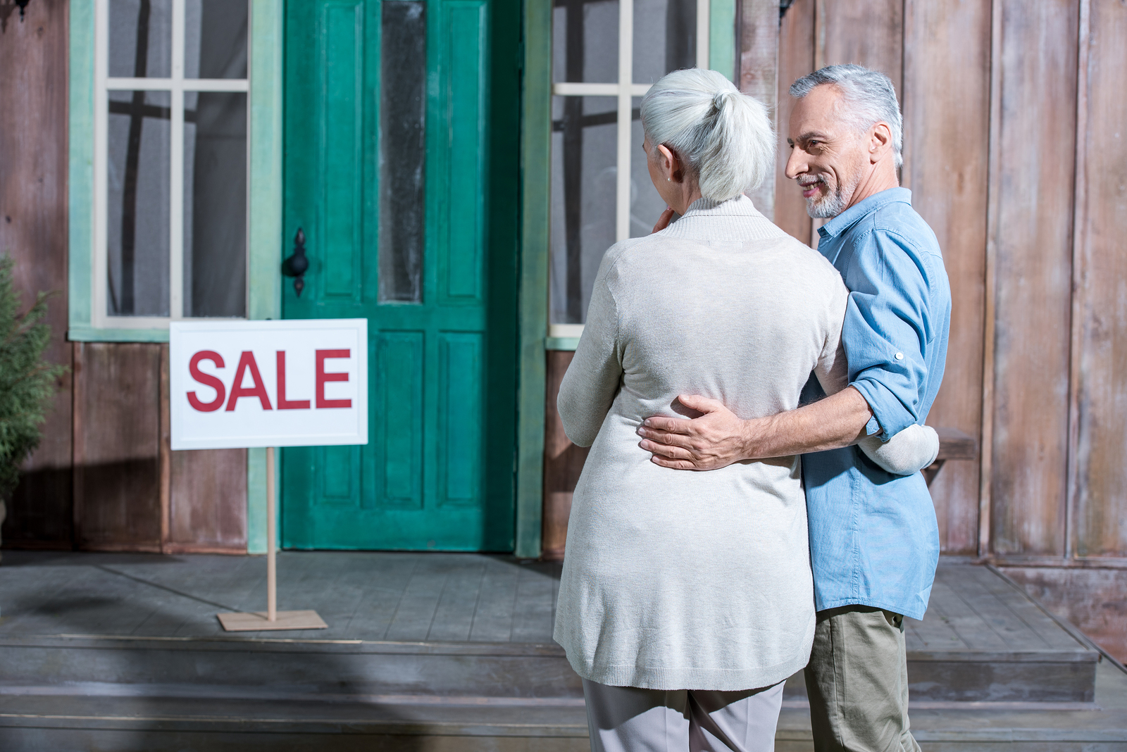 A senior woman and a senior man embrace side by side while looking at a sales sign in front of a house