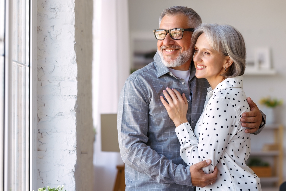 A senior couple embrace and smile for a photo while looking out a window.