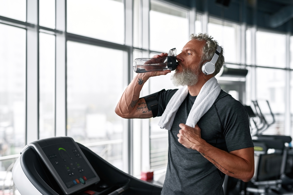 An older man takes a break from running on the treadmill to drink from his water bottle