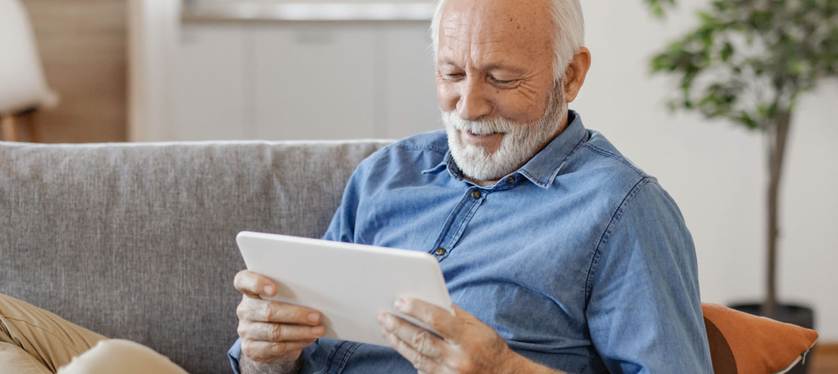 A senior man sitting on a couch while using a tablet.