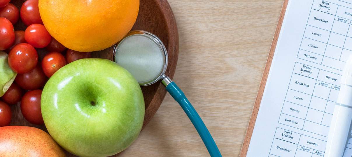 close up of a stethoscope, veggies, fruit, and a blood sugar tracker